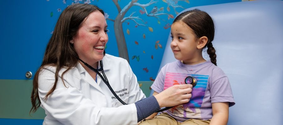 Physician Assistant Peggy Malloy in her white coat uses a stethoscope to examine a smiling young child sitting on an exam table in a brightly decorated pediatric clinic.