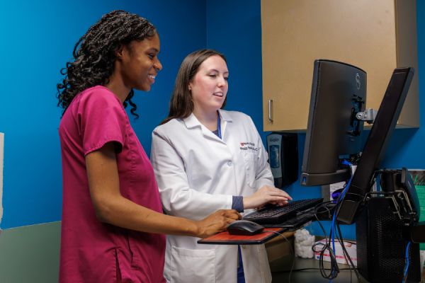Physician Assistant Peggy Malloy in her white coat and her coworker in pink scrubs stand at a computer station, discussing information on the monitor in a medical exam room.